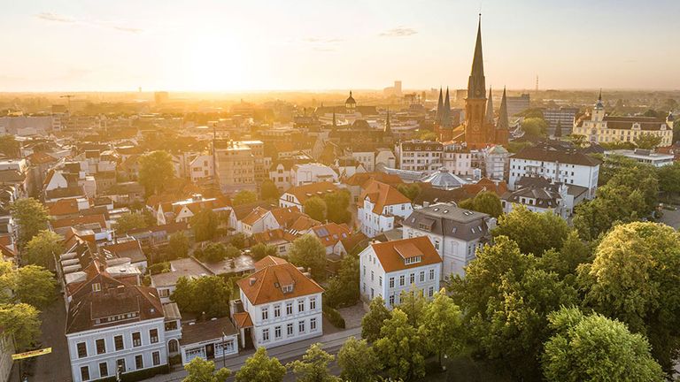 Alt-Text: „Sonnenuntergang über historische Altstadt mit Kirchtürmen und roten Dächern.“